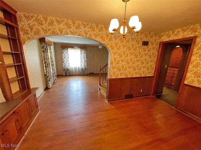 unfurnished dining area featuring wallpapered walls, arched walkways, a wainscoted wall, wood finished floors, and stairs