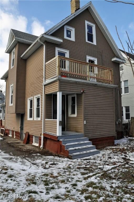 snow covered rear of property featuring a chimney and a balcony