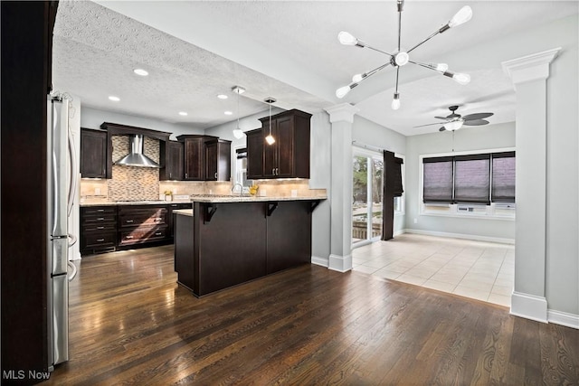 kitchen featuring dark brown cabinetry, a kitchen breakfast bar, wall chimney exhaust hood, tasteful backsplash, and ornate columns