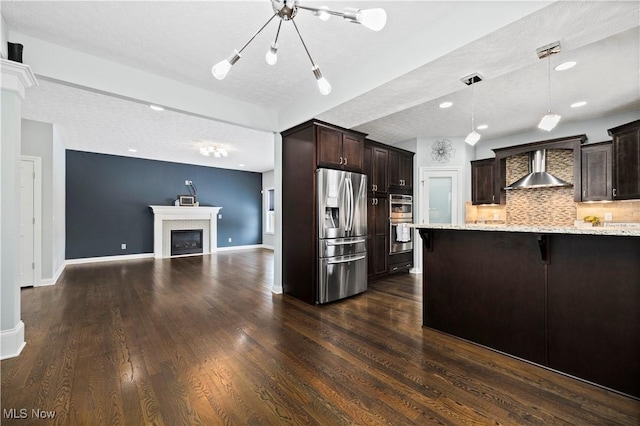 kitchen with dark brown cabinetry, decorative backsplash, stainless steel appliances, wall chimney range hood, and a fireplace