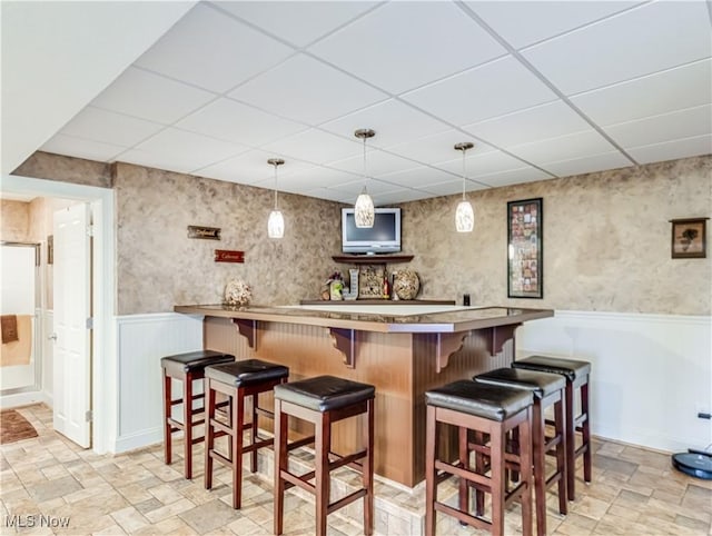 bar featuring a paneled ceiling, stone finish flooring, wainscoting, and bar