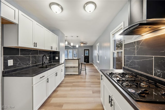 kitchen with dark countertops, wall chimney exhaust hood, light wood-style flooring, a sink, and black gas stovetop