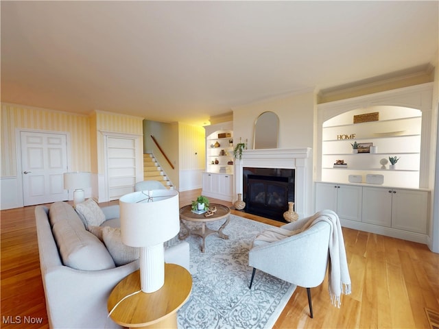 living area featuring built in shelves, stairs, a glass covered fireplace, light wood-type flooring, and wallpapered walls