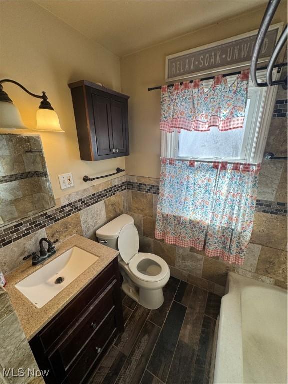 full bath with toilet, wood finished floors, vanity, tile walls, and wainscoting