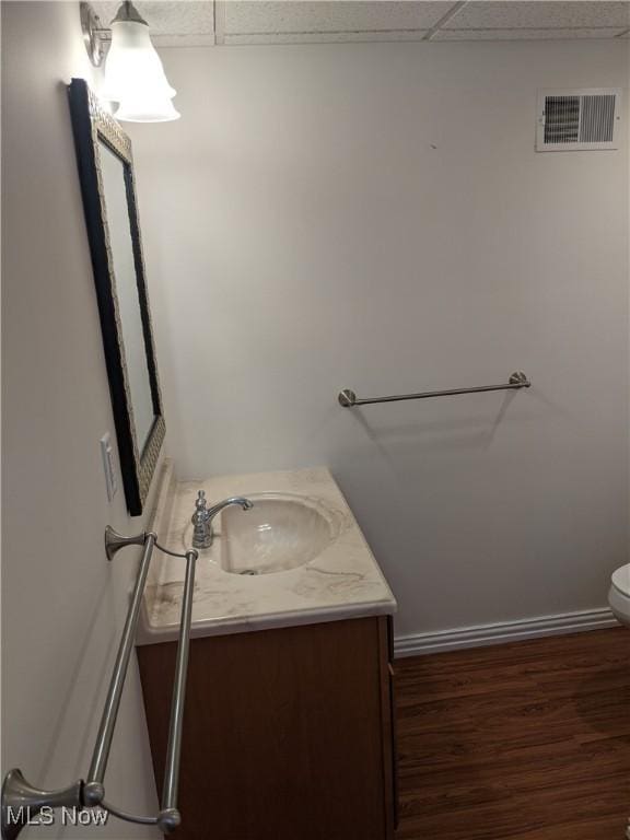 half bath featuring a drop ceiling, toilet, wood finished floors, vanity, and visible vents