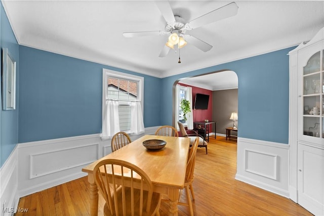 dining area with arched walkways, wainscoting, ceiling fan, and light wood-type flooring