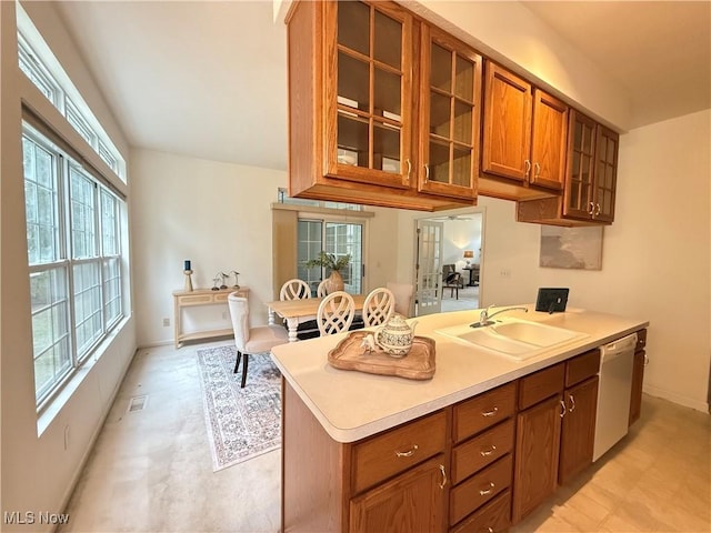 kitchen with brown cabinets, light countertops, stainless steel dishwasher, glass insert cabinets, and a sink