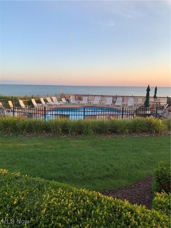 pool at dusk with a water view, a yard, and a community pool