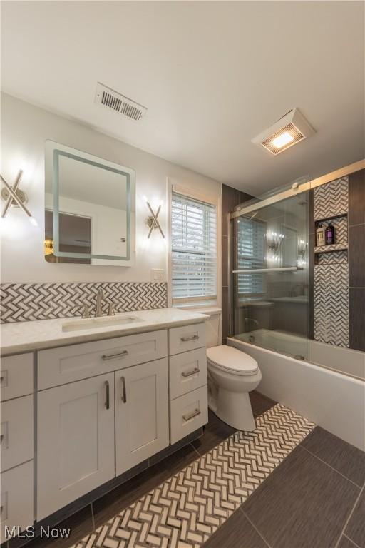 full bathroom with visible vents, bath / shower combo with glass door, decorative backsplash, toilet, and vanity