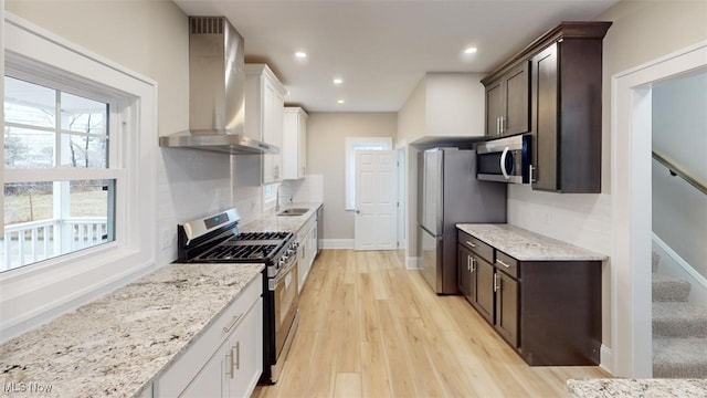 kitchen with dark brown cabinetry, light wood finished floors, wall chimney exhaust hood, stainless steel appliances, and backsplash