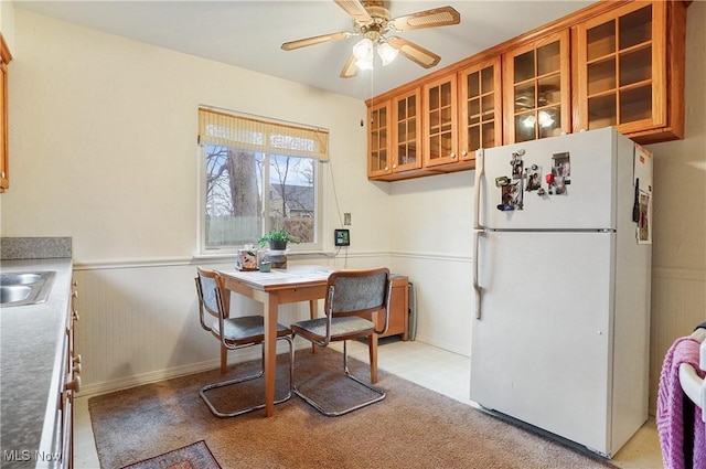 kitchen featuring ceiling fan, wainscoting, freestanding refrigerator, brown cabinets, and glass insert cabinets