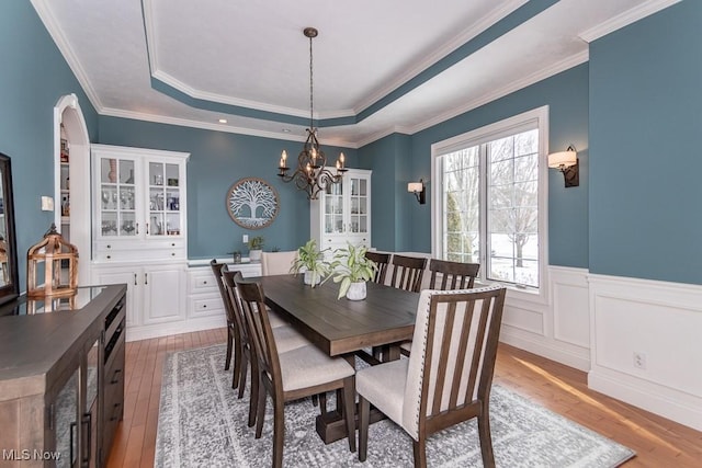 dining area with a raised ceiling, wainscoting, light wood-style flooring, ornamental molding, and a notable chandelier