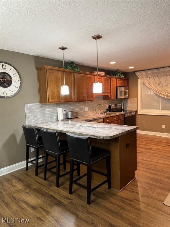 kitchen with dark wood-style floors, backsplash, appliances with stainless steel finishes, brown cabinetry, and a peninsula