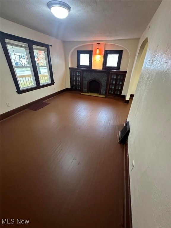 unfurnished living room featuring arched walkways, a fireplace with raised hearth, a textured ceiling, wood finished floors, and baseboards