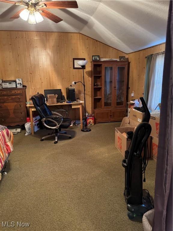 carpeted home office with lofted ceiling, ceiling fan, and wood walls