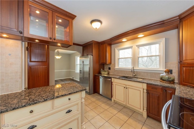 kitchen featuring stainless steel appliances, a sink, cream cabinetry, dark stone counters, and glass insert cabinets