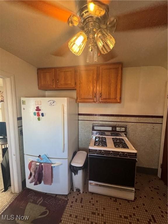 kitchen with brown cabinets, tile walls, a ceiling fan, wainscoting, and white appliances