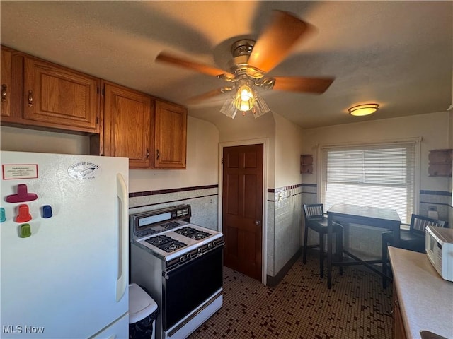 kitchen featuring a wainscoted wall, white appliances, a ceiling fan, tile walls, and brown cabinetry