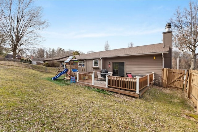 back of property featuring a deck, a playground, fence, a lawn, and a chimney