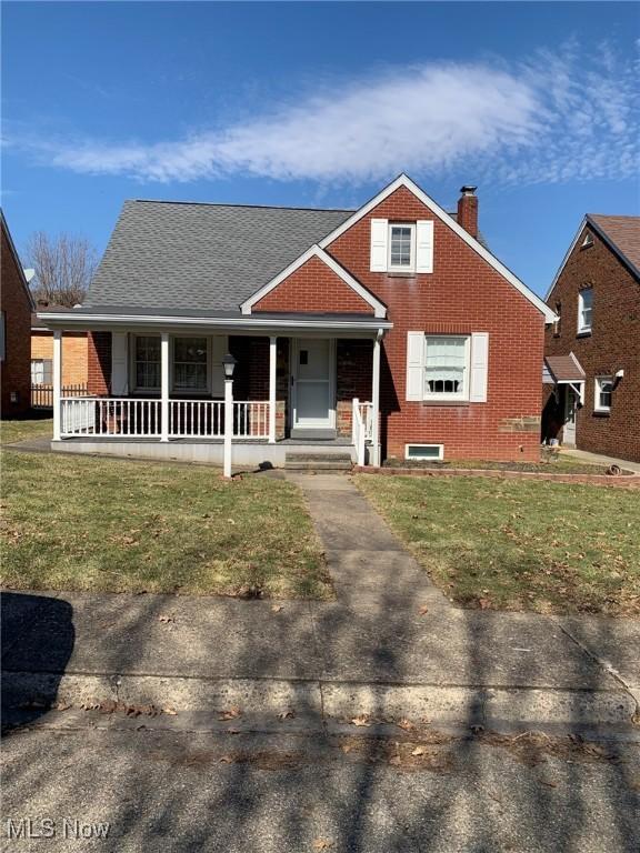 view of front facade featuring brick siding, roof with shingles, a chimney, covered porch, and a front yard