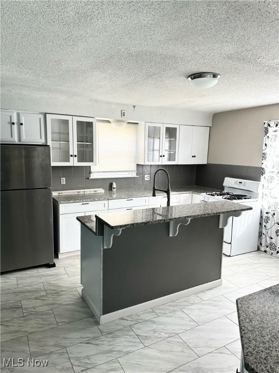 kitchen featuring gas range gas stove, a breakfast bar area, freestanding refrigerator, a kitchen island with sink, and white cabinetry