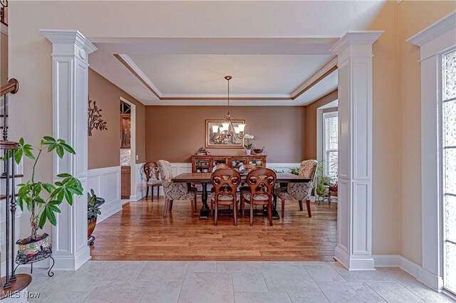 dining room featuring a chandelier, light wood-type flooring, wainscoting, a tray ceiling, and ornate columns