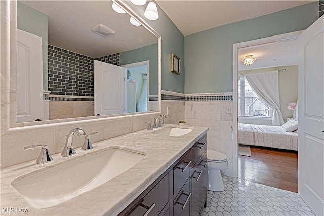 ensuite bathroom featuring toilet, double vanity, a sink, and tile walls