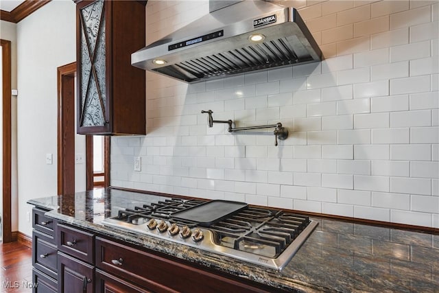 kitchen with dark stone counters, dark wood finished floors, wall chimney range hood, stainless steel gas stovetop, and backsplash