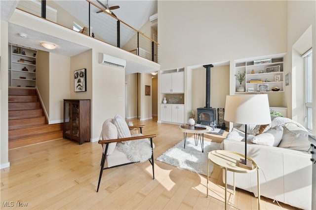 living area with a towering ceiling, a wall mounted air conditioner, a wood stove, stairs, and light wood-style floors