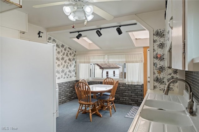 dining room with wallpapered walls, a ceiling fan, a wainscoted wall, brick wall, and rail lighting