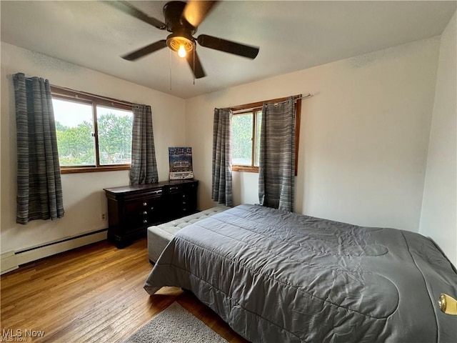 bedroom with a baseboard heating unit, multiple windows, wood finished floors, and a ceiling fan