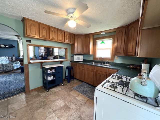 kitchen featuring arched walkways, dark countertops, brown cabinetry, a sink, and white range with gas stovetop