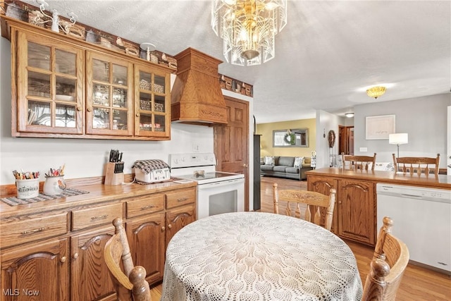 kitchen featuring white appliances, light wood finished floors, glass insert cabinets, custom exhaust hood, and a chandelier