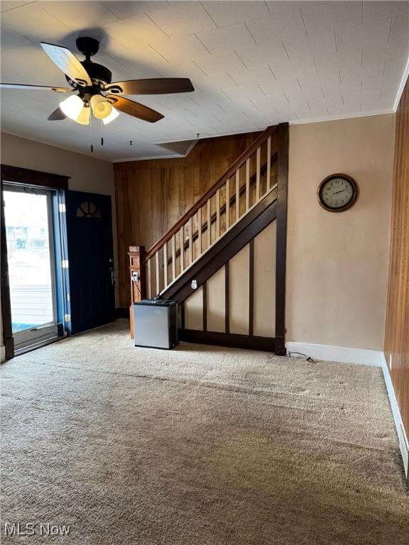 carpeted foyer featuring a ceiling fan, ornamental molding, wooden walls, baseboards, and stairs