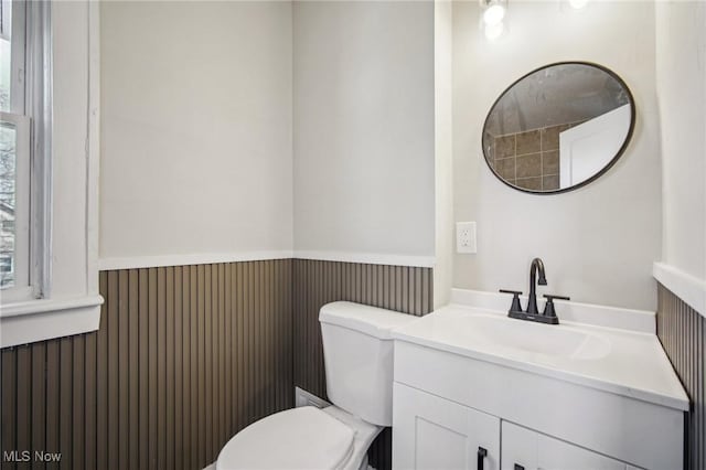 half bath with toilet, plenty of natural light, vanity, and a wainscoted wall