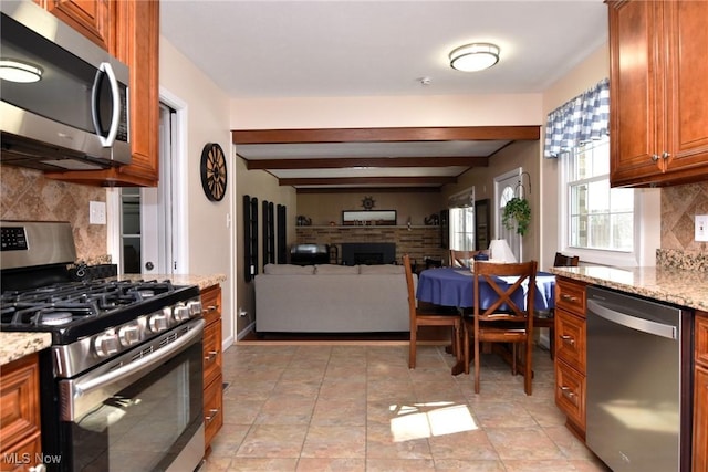 kitchen with light stone counters, tasteful backsplash, appliances with stainless steel finishes, brown cabinetry, and beamed ceiling