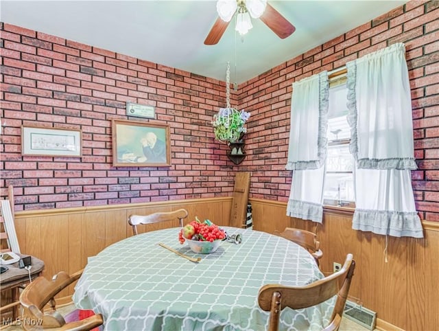 dining space featuring a ceiling fan, wainscoting, wooden walls, and brick wall