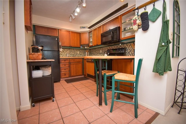 kitchen with light tile patterned floors, brown cabinets, decorative backsplash, black appliances, and glass insert cabinets