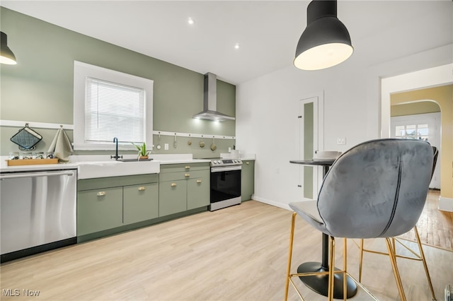 kitchen with light wood finished floors, stainless steel appliances, green cabinets, a sink, and wall chimney range hood