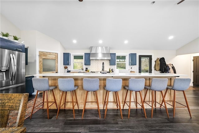 kitchen featuring lofted ceiling, light countertops, blue cabinetry, stainless steel fridge with ice dispenser, and wall chimney exhaust hood