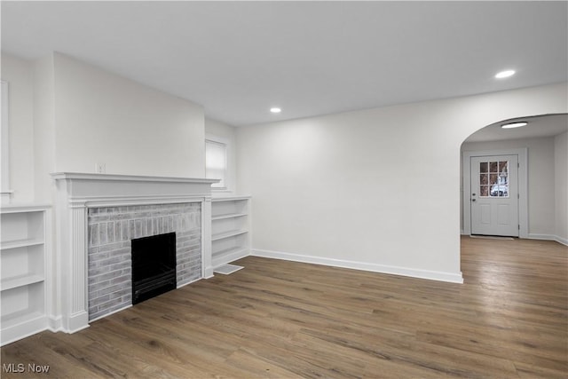 unfurnished living room with arched walkways, recessed lighting, a brick fireplace, wood finished floors, and baseboards