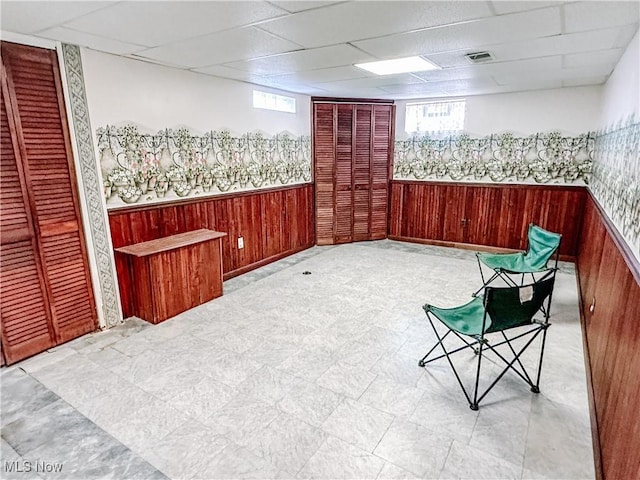 sitting room featuring a wainscoted wall, wood walls, a drop ceiling, and a healthy amount of sunlight