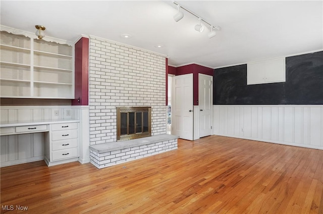 unfurnished living room with a brick fireplace, light wood-style flooring, built in desk, and a wainscoted wall