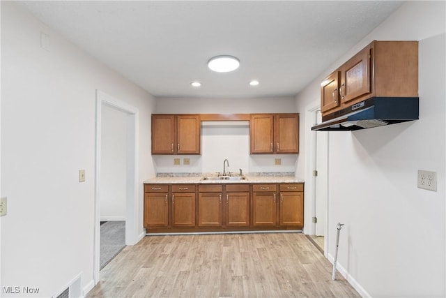 kitchen with light wood finished floors, brown cabinetry, light countertops, under cabinet range hood, and a sink