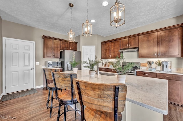 kitchen featuring a center island with sink, stainless steel appliances, a sink, wood finished floors, and under cabinet range hood