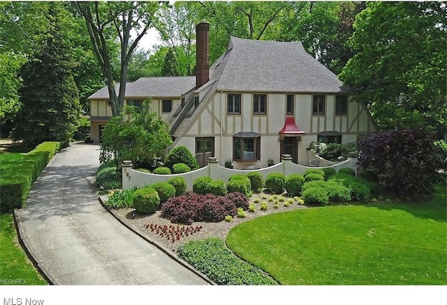 tudor house featuring a shingled roof, concrete driveway, stucco siding, a chimney, and a front yard