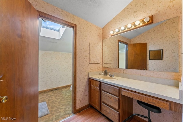 bathroom with lofted ceiling with skylight, vanity, wood finished floors, baseboards, and wallpapered walls