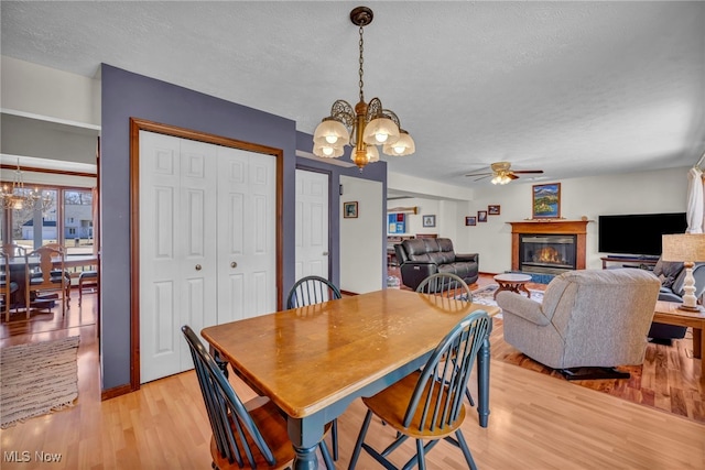 dining room featuring a textured ceiling, light wood finished floors, ceiling fan with notable chandelier, and a glass covered fireplace