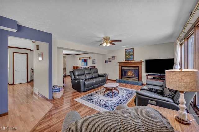 living area featuring light wood-style flooring, a ceiling fan, a glass covered fireplace, a textured ceiling, and baseboards