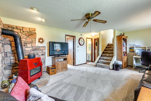 living room featuring light colored carpet, stairway, a ceiling fan, a wood stove, and a textured ceiling